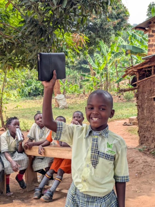 A smiling pupil holding a book above his head, classmates on a bench behind him