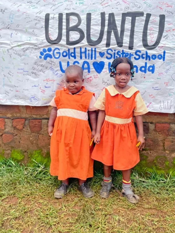 Two young pupils in orange uniforms standing in front of a banner signed by Gold Hill Sister School